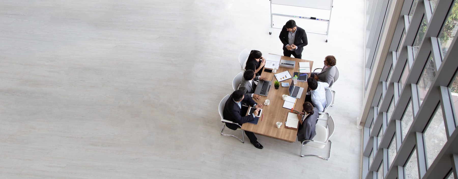 Aerial view of a business meeting. Six people at a wooden table with laptops, papers, and a plant. Large windows on the right.