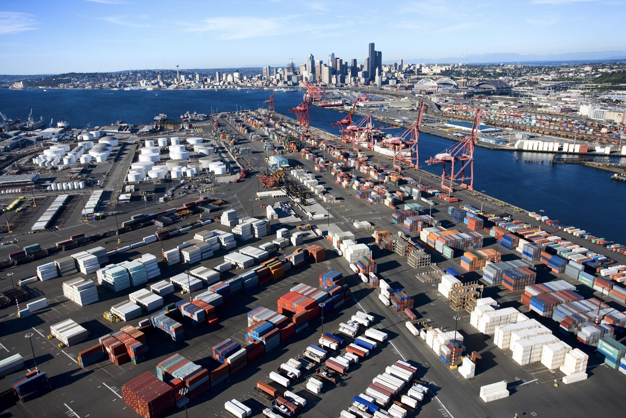 Aerial view of a busy port with colorful shipping containers and red cranes. City skyline and blue water in the background under a clear sky.
