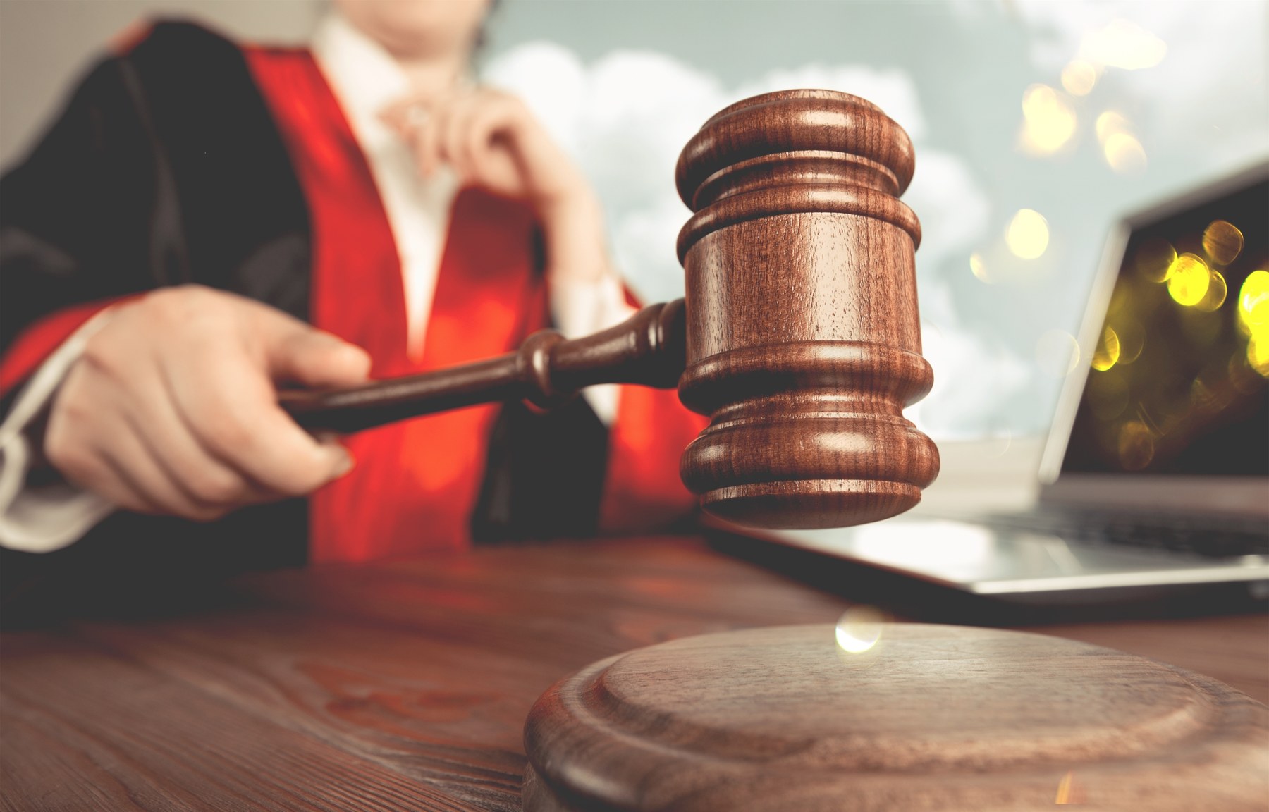 Judge in red and black robes motions with gavel over wooden desk. A laptop is nearby. Bright, warm light suggests a serious setting.