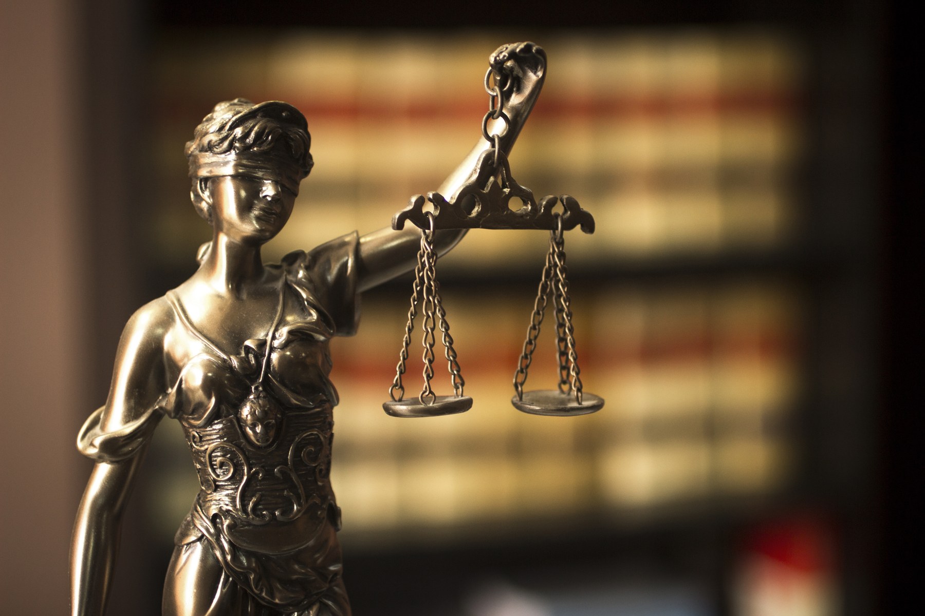 Bronze Lady Justice statue holding scales in a library setting; blurred books in the background. Dim lighting evokes solemnity.