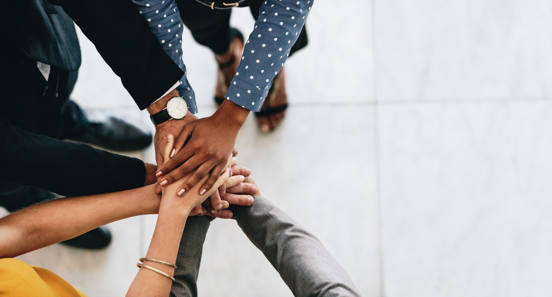 Hands of diverse people stacked in teamwork over a light tiled floor. Individuals wear various patterned sleeves, watch, and bracelets.