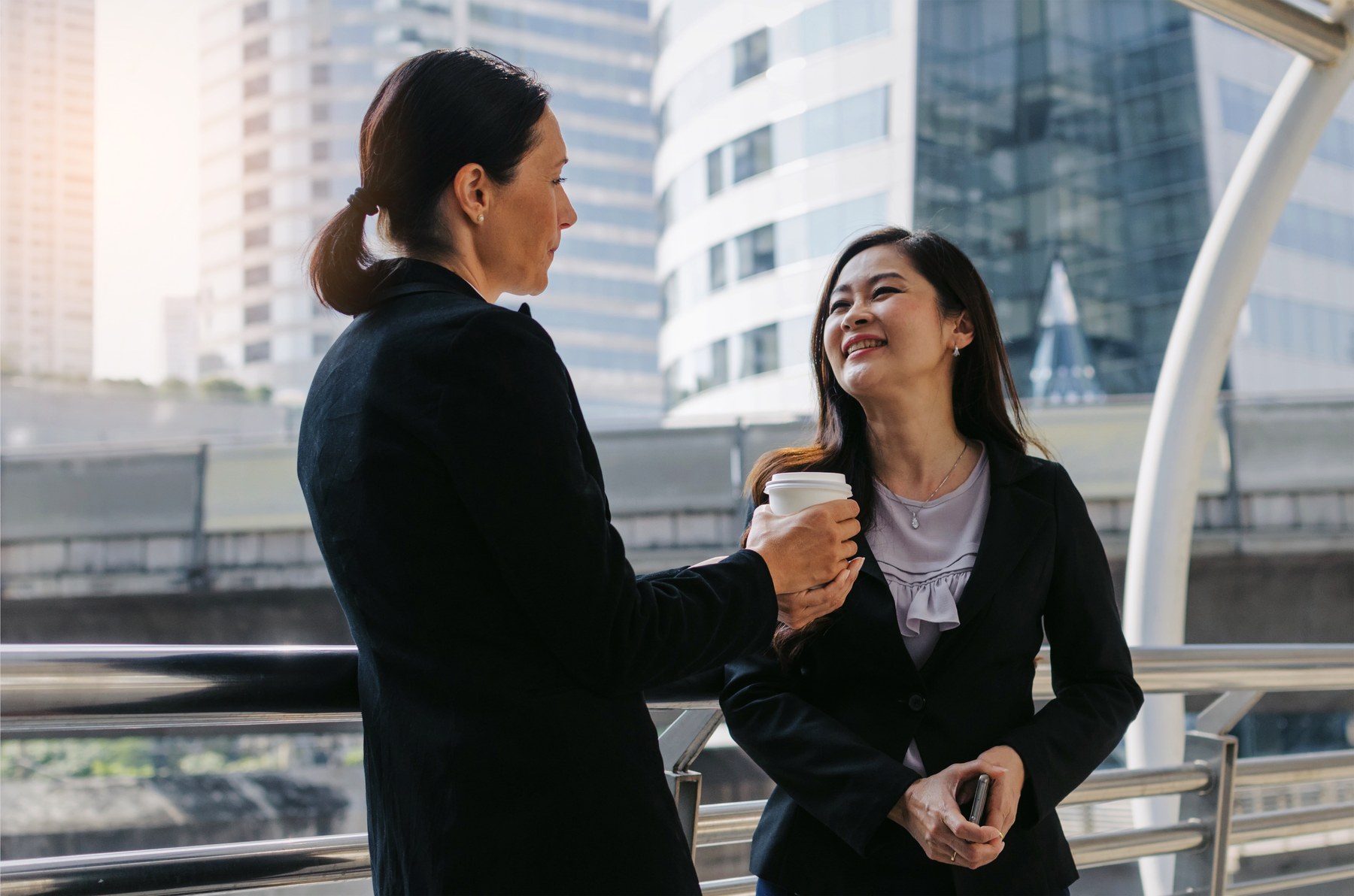 Two women in business attire converse outdoors, one holding a coffee cup. Background shows modern skyscrapers. Both appear engaged and smiling.