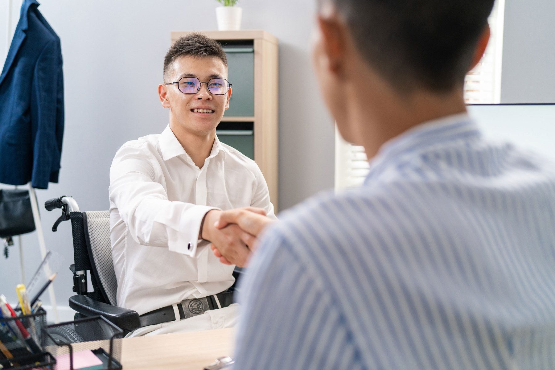 Two people in an office shaking hands. One is in a wheelchair, smiling, wearing glasses, and a white shirt. A blazer hangs in the background.