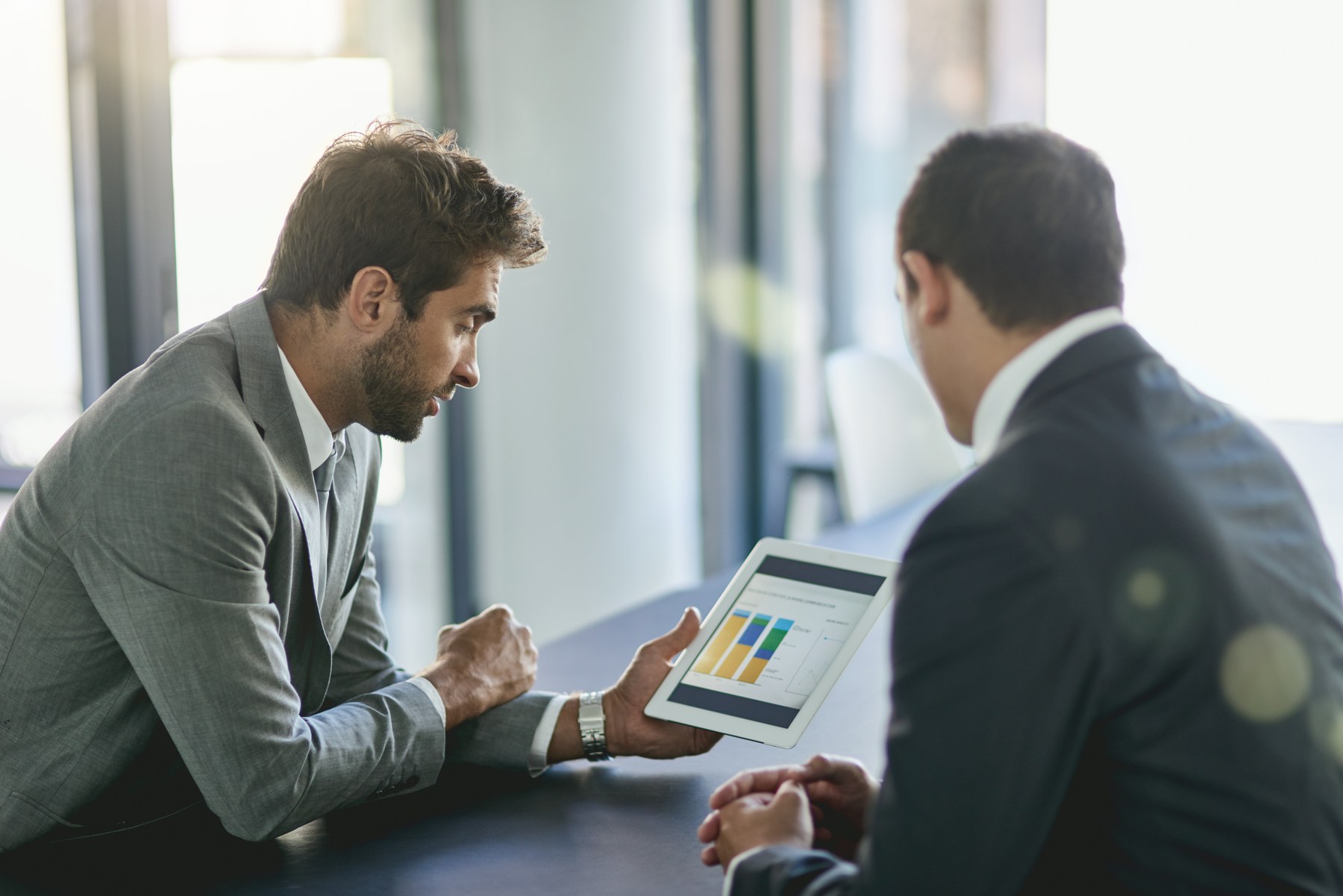 Two men in suits discuss a tablet displaying colorful graphs in a bright office setting. One looks engaged, sunlight filters in.