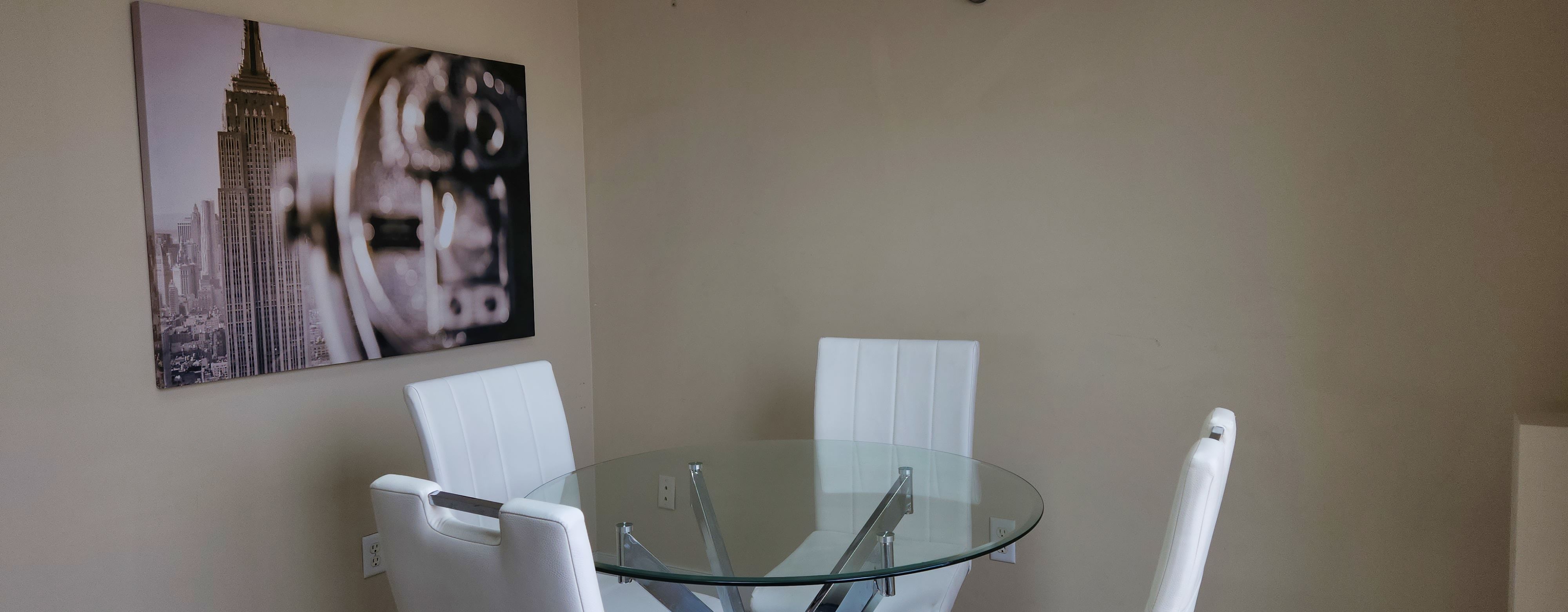 Modern dining area with a round glass table and four white upholstered chairs, and a black-and-white photo of the Empire State Building on the wall. Flex Stay Corporate Bella Vista Penthouse, Calgary.