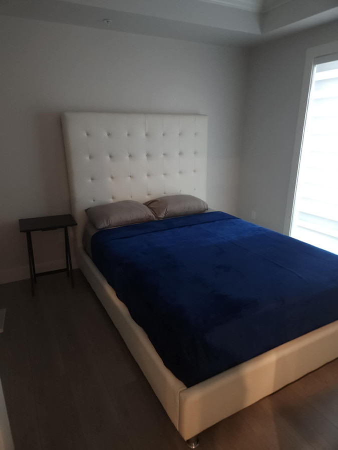 Minimalist bedroom with a white tufted headboard bed, navy blue blanket, two gray pillows, and a small dark wooden side table. Oak Street Signature Suite: High-Rise Luxury in Marpole