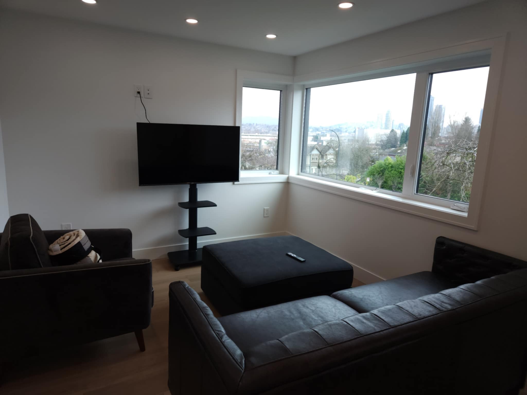 Modern living room with black leather sofa, black armchair holding a folded blanket, black ottoman with remote control, wall-mounted flat-screen TV on stand, and large windows showing a cityscape view. Rupert Street Executive Retreat: Proximity & Peace