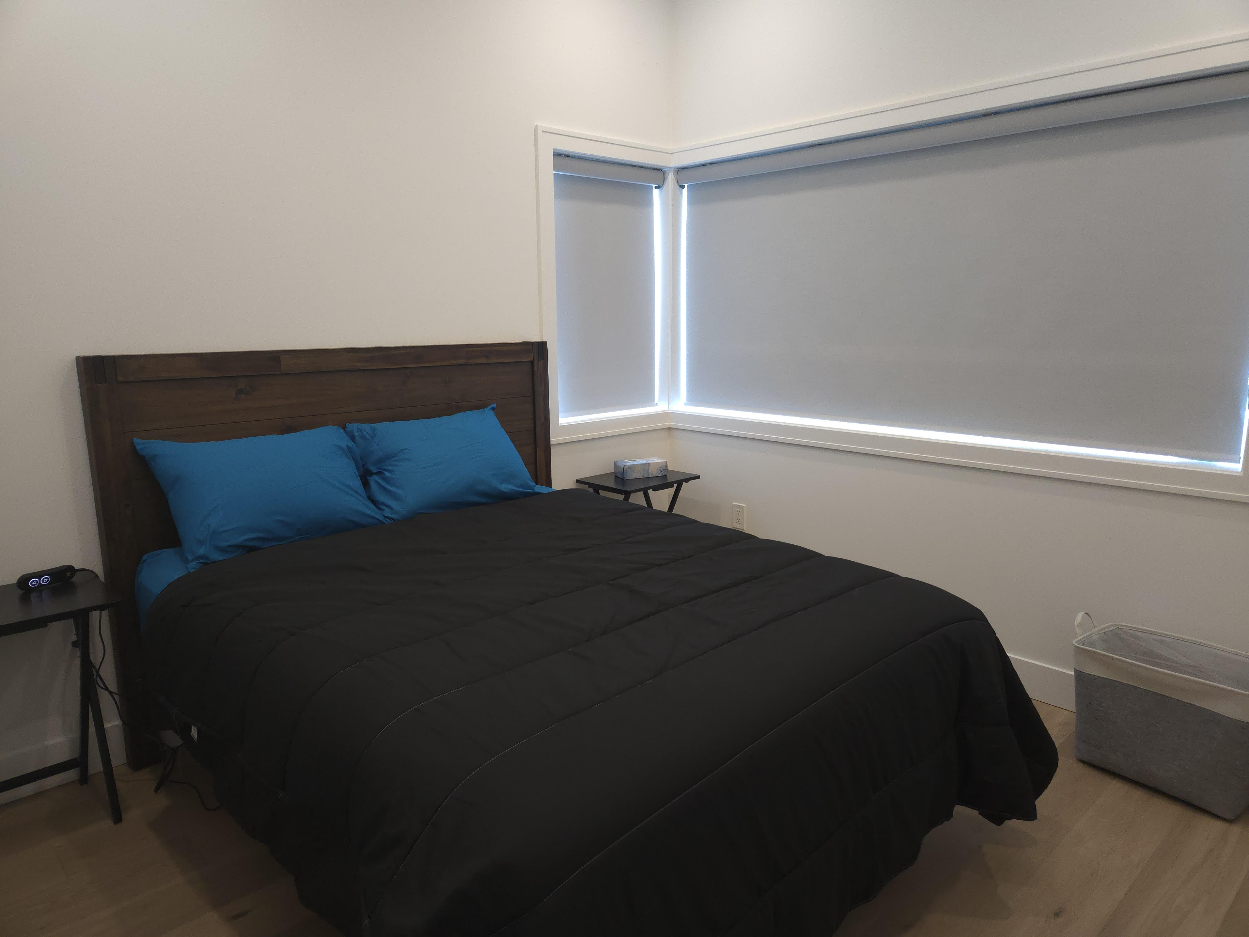Bedroom with a wooden headboard bed made with black bedding and blue pillows, two small black side tables, white walls, and windows with closed gray blinds. Rupert Street Executive Retreat: Proximity & Peace