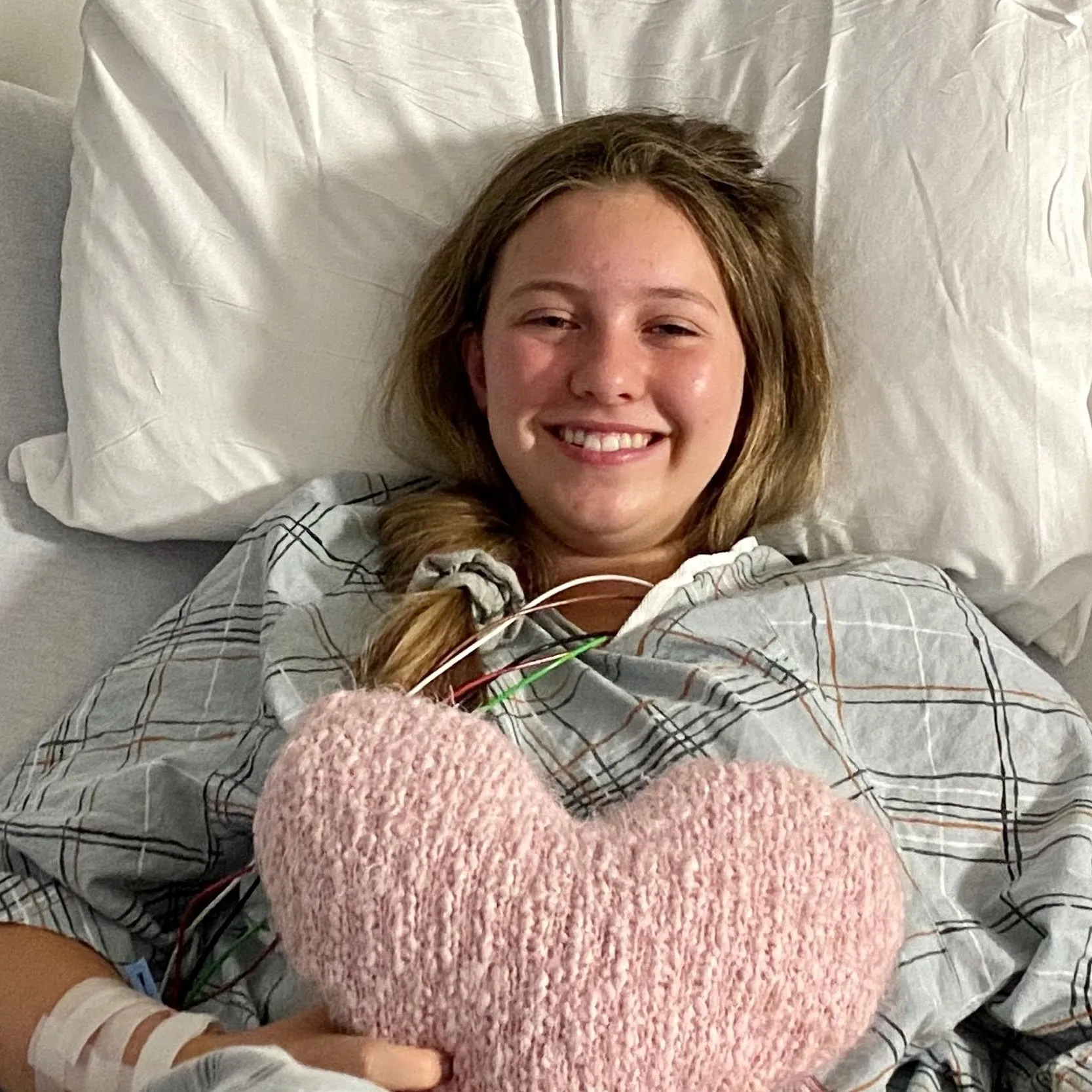 Young woman smiling in a hospital bed while holding a heart-shaped pillow