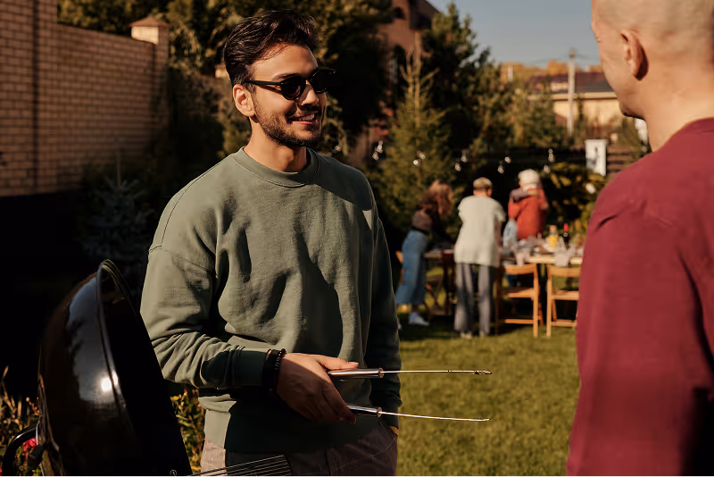 person in a green jumper holding tongs standing over a barbecue chatting to another person in a garden