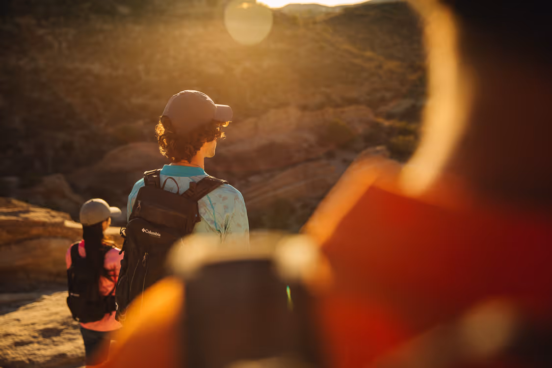 Two people hiking outdoors with backpacks and caps, bathed in warm sunlight near rocky terrain.