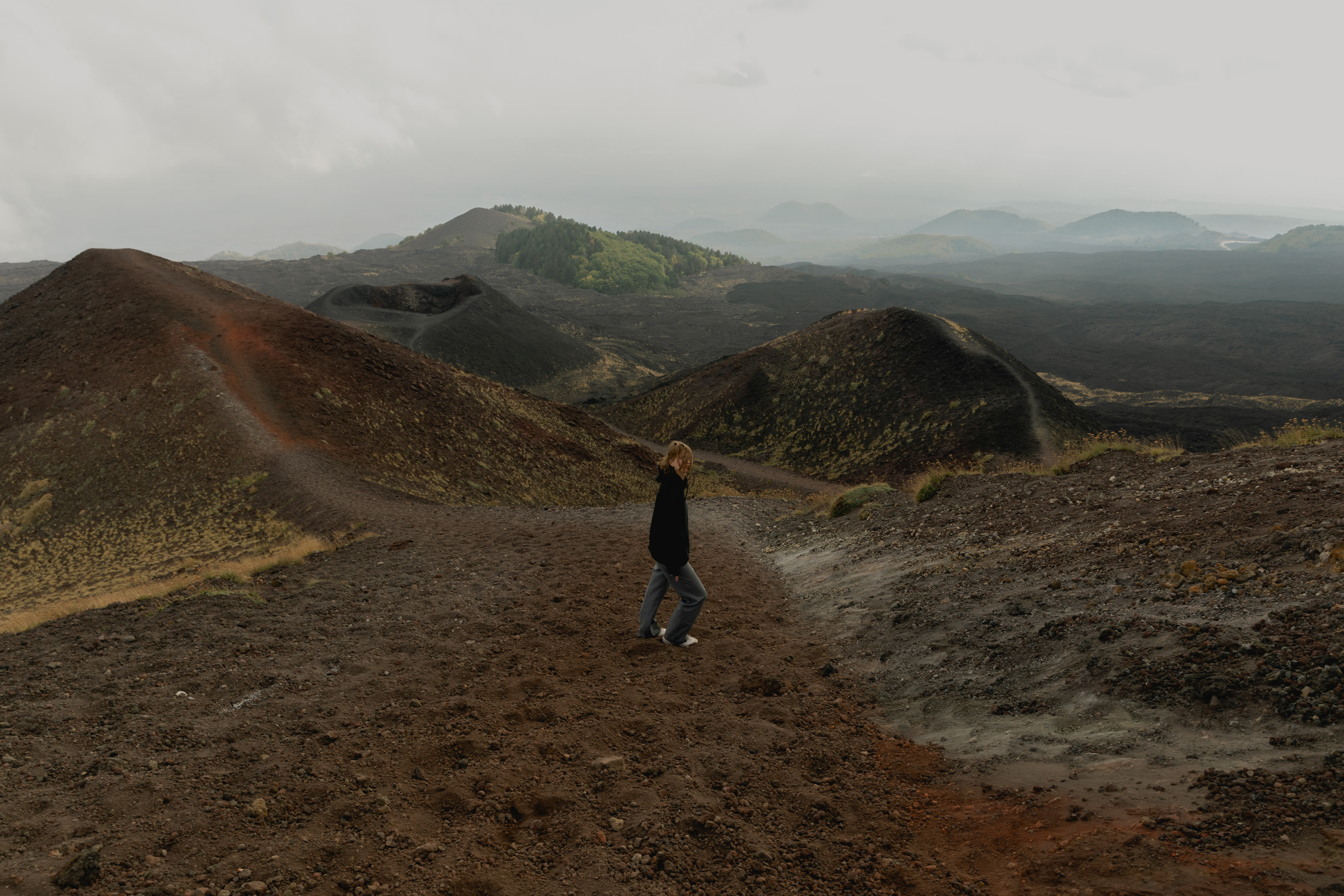 Person walking on volcanic terrain with dark hills and green patches under a cloudy sky.