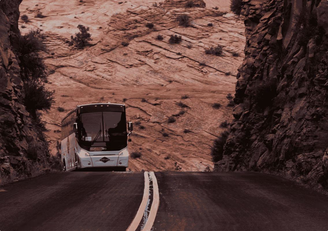 White bus driving on road between rocky cliffs with desert terrain in the background.