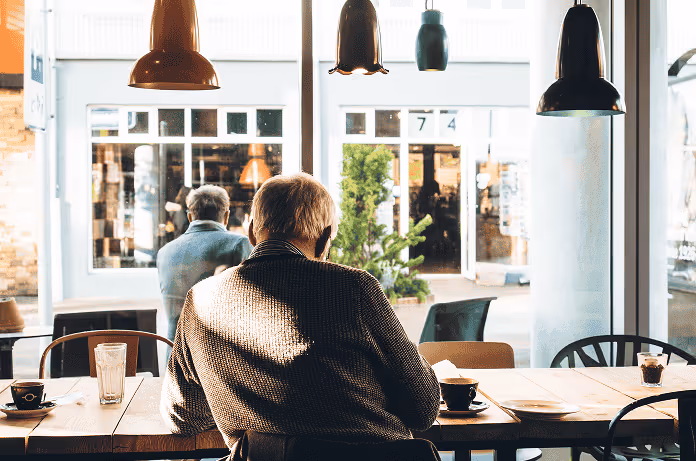 Man sitting alone at a wooden table in a cafe with coffee cups and glasses near large windows.