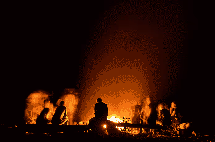 Group of people sitting around a glowing campfire at night with dark background.