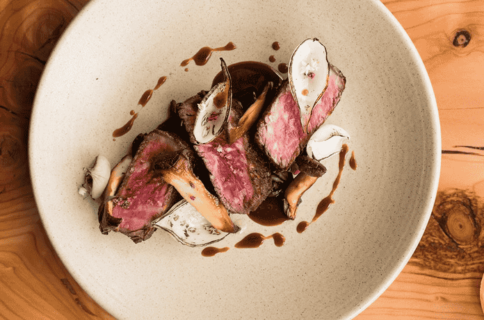 Slices of medium-rare steak with mushrooms and sauce served on a white plate atop a wooden table.