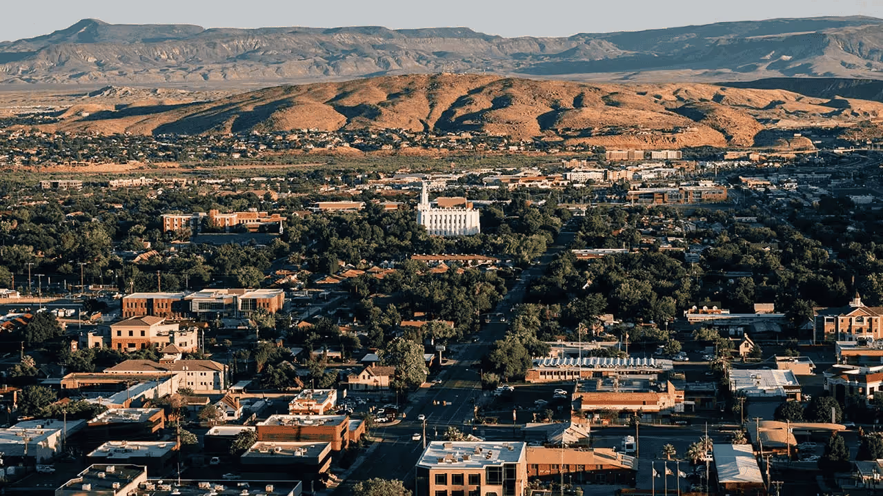 Cityscape with clustered buildings and trees, desert hills and mountains in the background at sunset.