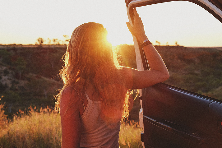 Person with long hair standing by an open car door watching the sunset in a grassy landscape.