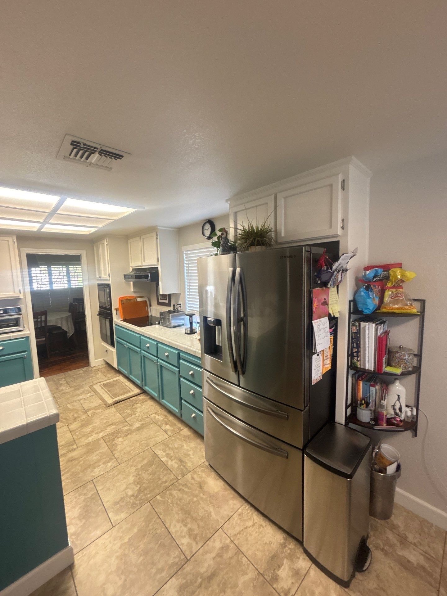 Kitchen with teal lower cabinets, white upper cabinets, stainless steel French door refrigerator, and a small shelf with snacks and books next to the fridge.