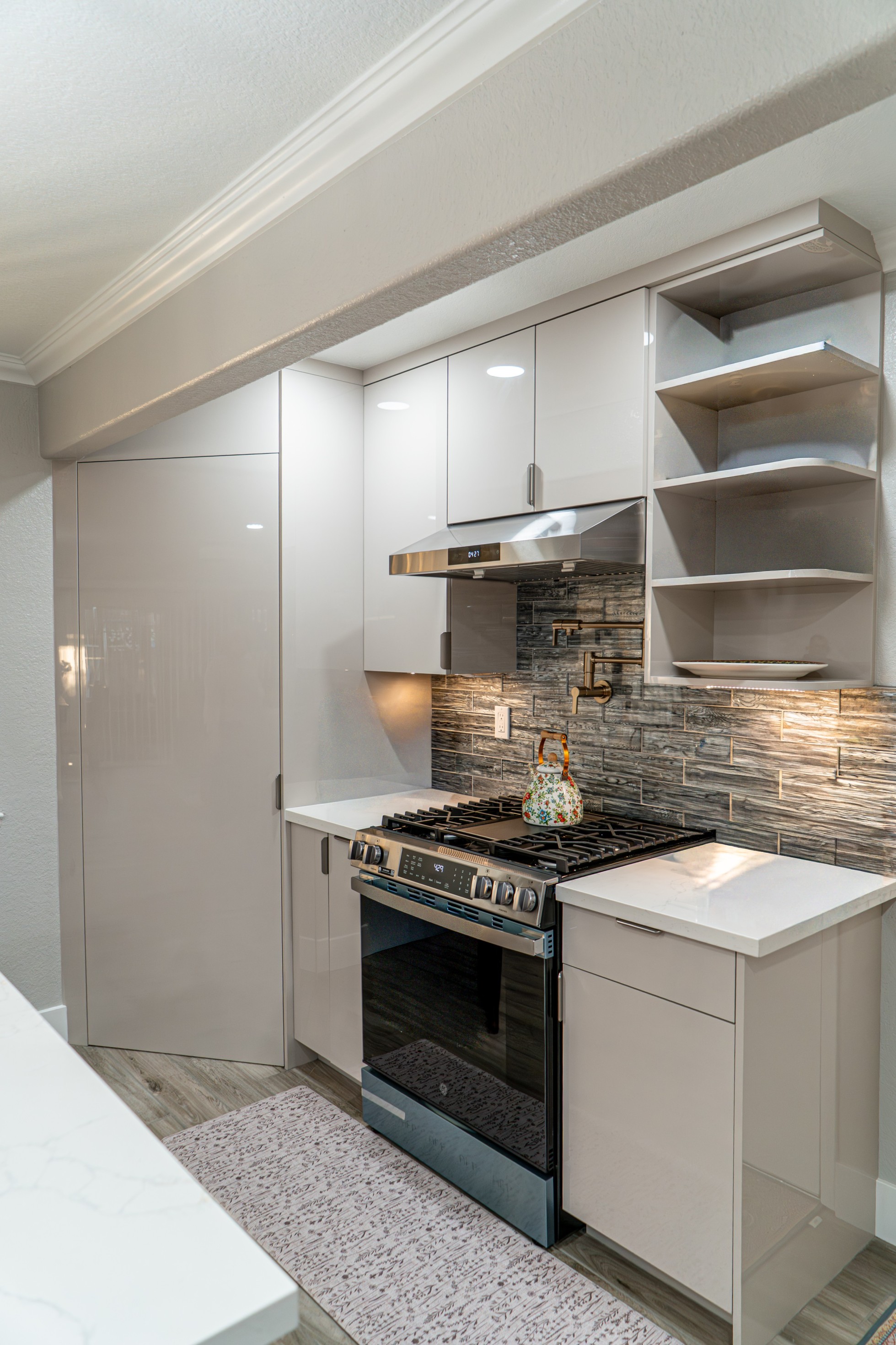 Modern kitchen corner with stainless steel gas stove, white glossy cabinets, textured gray backsplash, and a floral teapot on the stove.