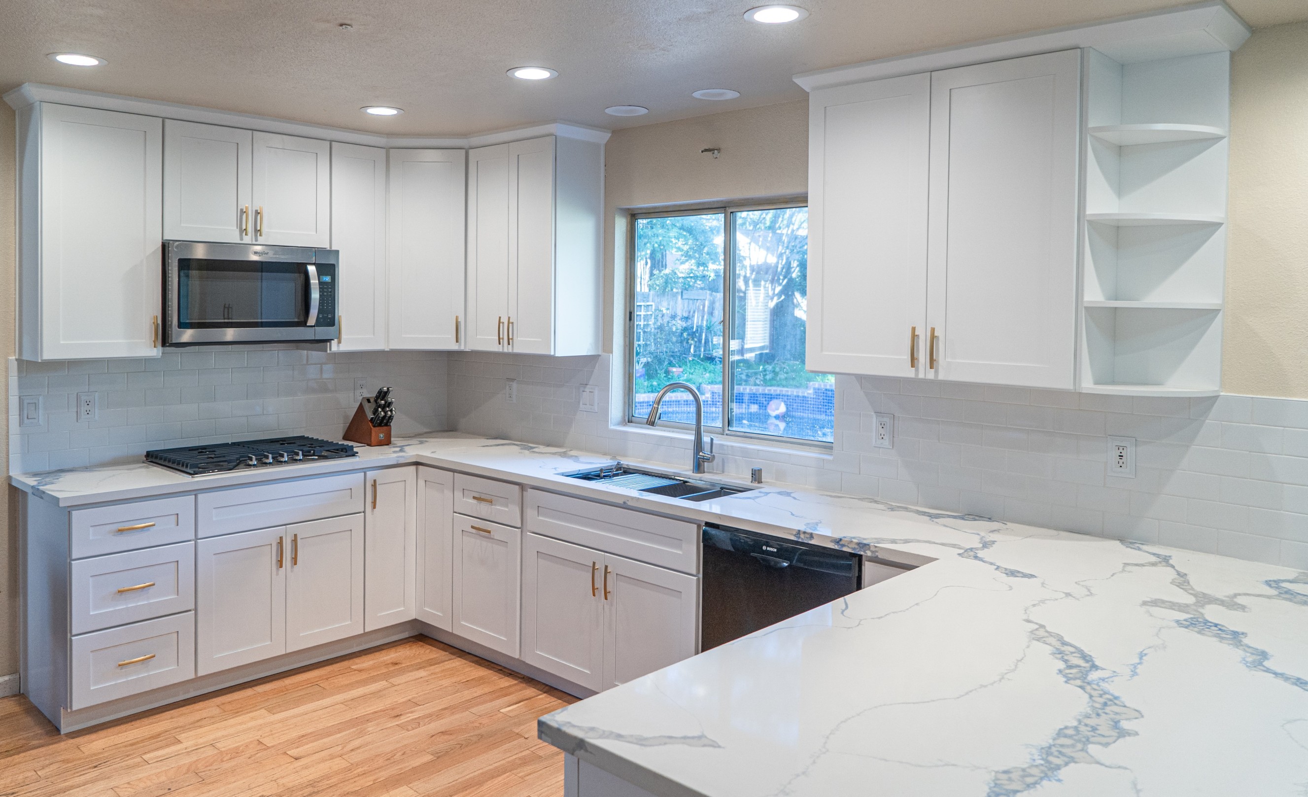 Modern kitchen with white cabinets, marble-patterned countertops, stainless steel microwave, gas stove, and a window above the sink.