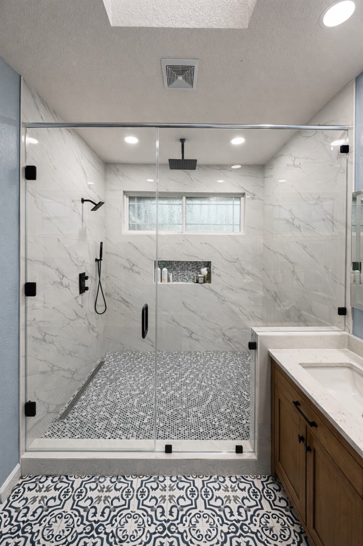 Modern bathroom shower with glass doors, marble walls, mosaic tile floor, and black rain showerhead and fixtures.