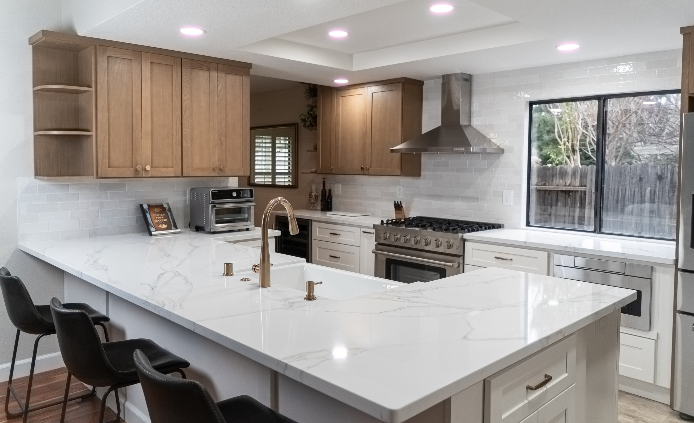 Modern kitchen with white marble countertops, wooden upper cabinets, stainless steel stove and hood, and black bar stools.