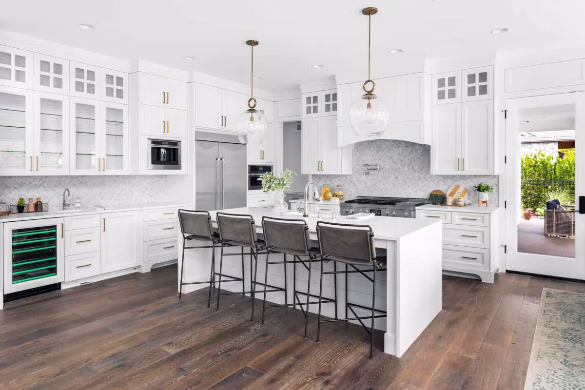 Modern white kitchen with large island, four bar stools, pendant lights, stainless steel appliances, and hardwood flooring.
