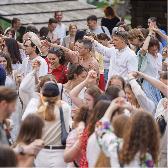 A large group of young people dancing closely together outdoors, many wearing traditional embroidered clothing.