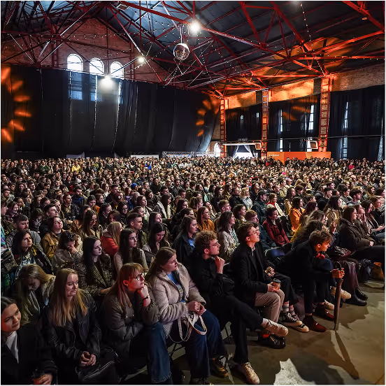 Large audience seated indoors in a spacious venue with red metal beams and arched windows, attentively watching a presentation or event.