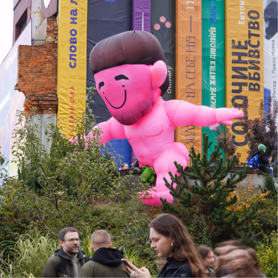 Large pink inflatable figure with brown hair and smiling face outdoors near colorful vertical banners, with people and greenery in the foreground.