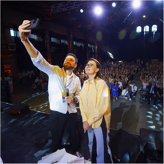Man and woman on stage taking a selfie with a smartphone in front of a cheering crowd in a large indoor venue.
