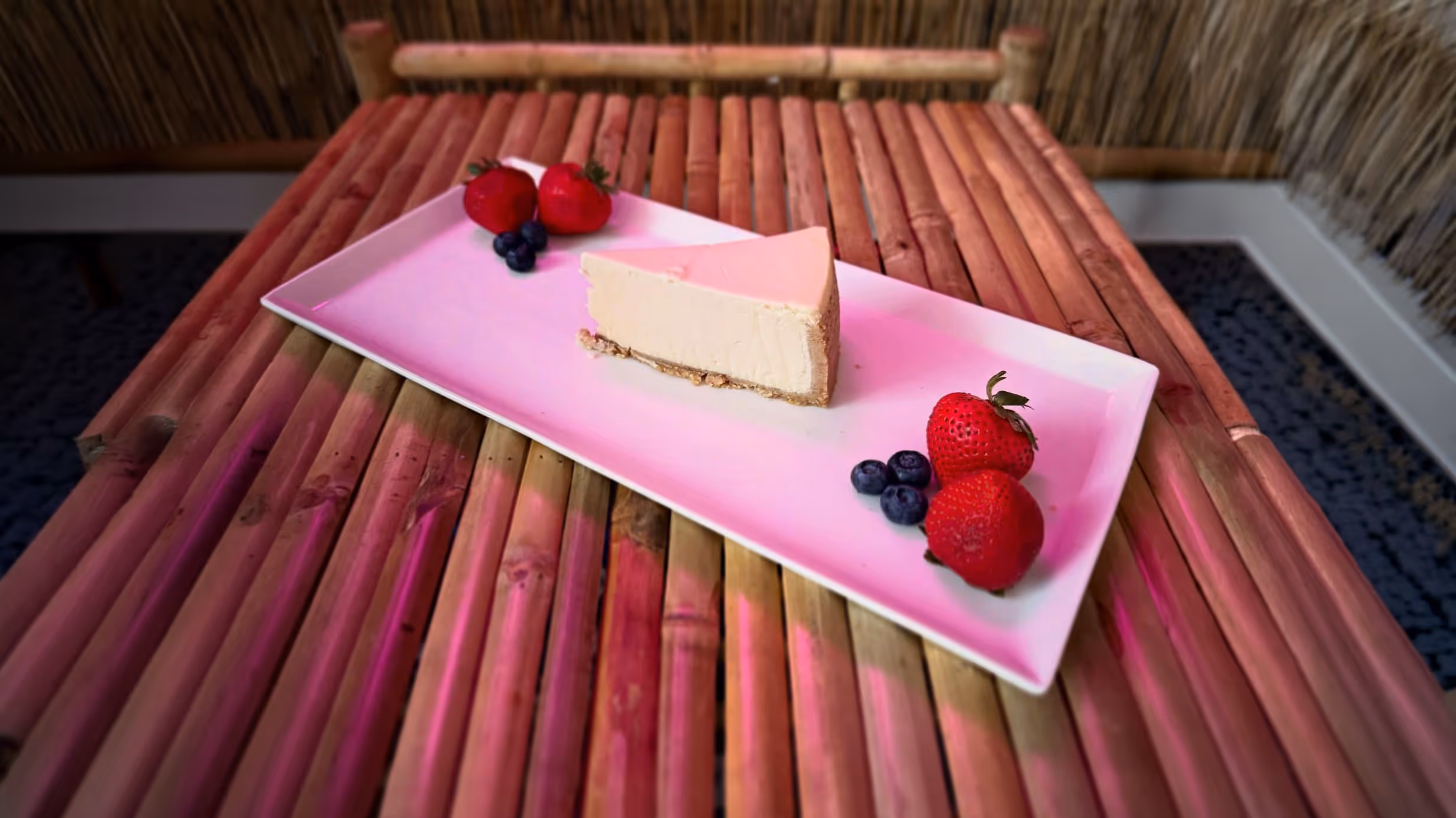 Slice of cheesecake on a rectangular white plate with strawberries and blueberries, placed on a bamboo table.