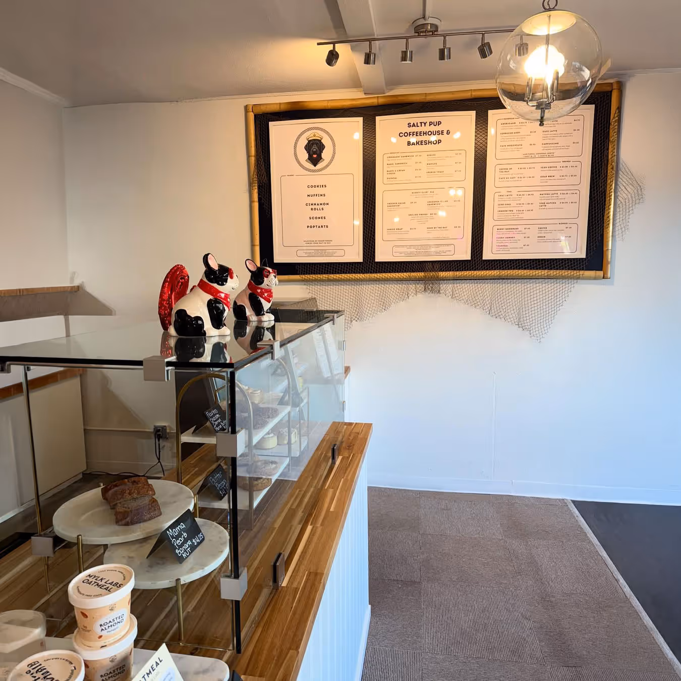 Interior of a coffeehouse with a wooden counter, glass display case holding pastries, two ceramic dog figurines with red bandanas, and a menu board on the wall.