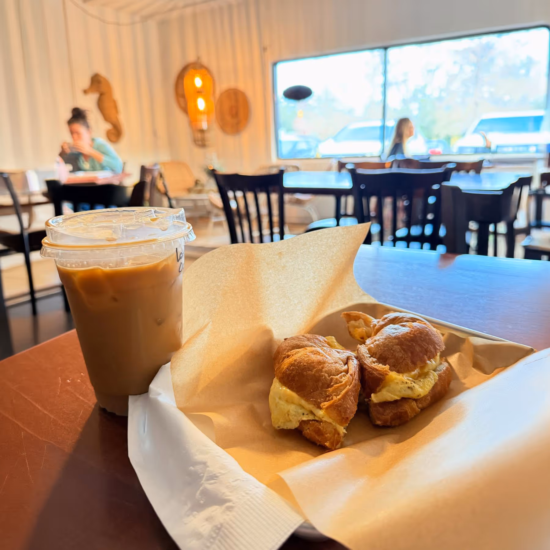 Iced coffee and a croissant breakfast sandwich with egg on a tray inside a cafe with people in the background.
