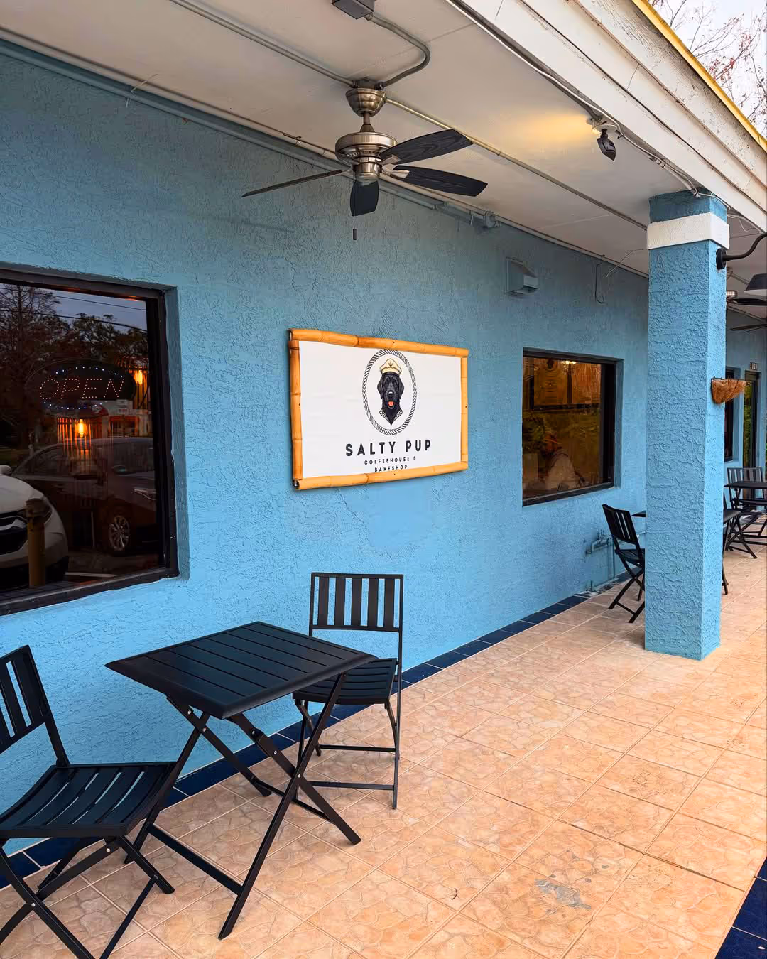Outdoor seating area with black tables and chairs outside a blue building with a 'Salty Pup Coffeehouse & Bakeshop' sign featuring a black dog wearing a sailor hat.