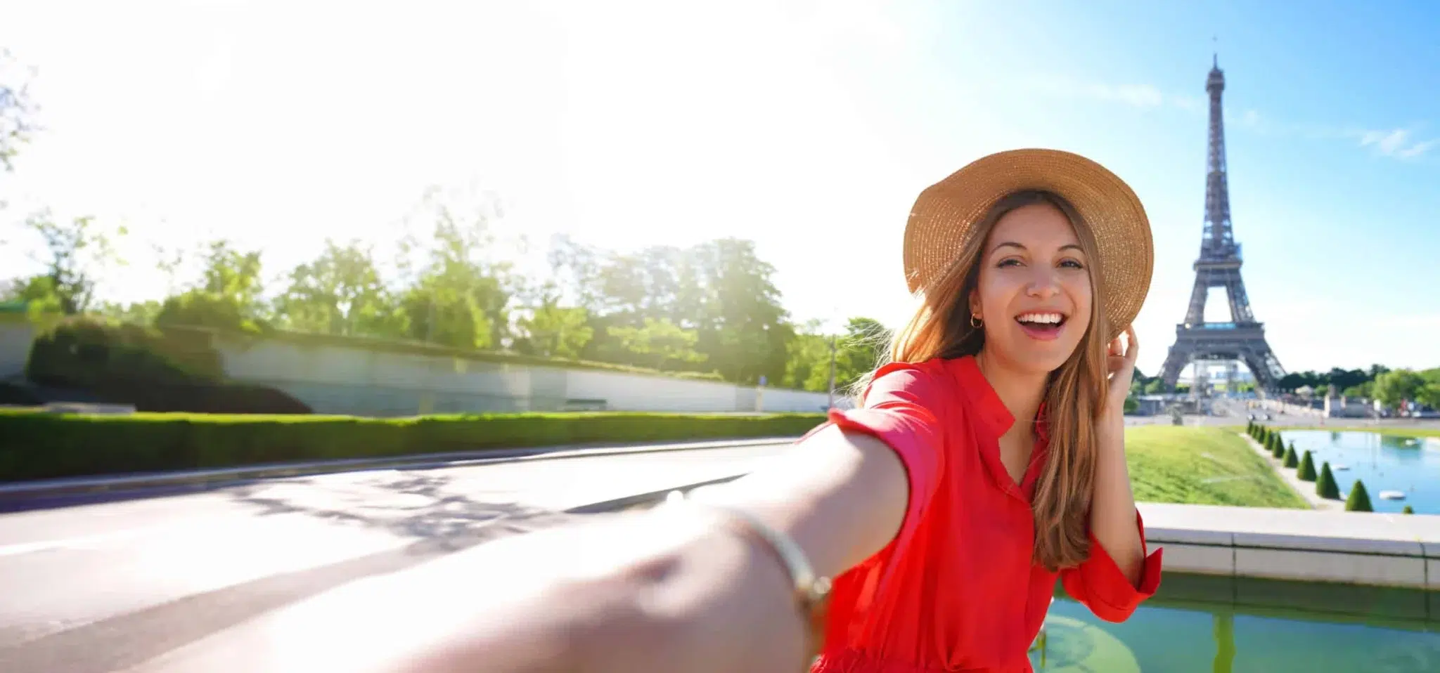 Woman in a red dress and hat taking a selfie with Eiffel Tower in the background on sunny day.