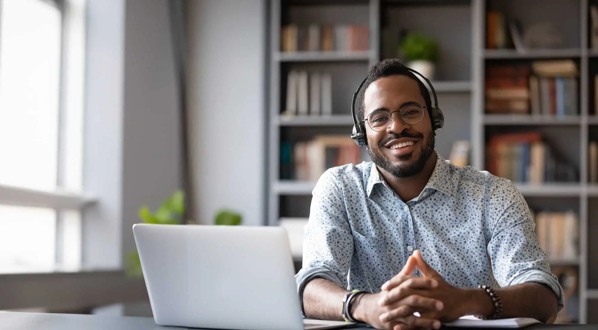 Smiling man wearing glasses and headset sitting at a desk with a laptop in a room with bookshelves
