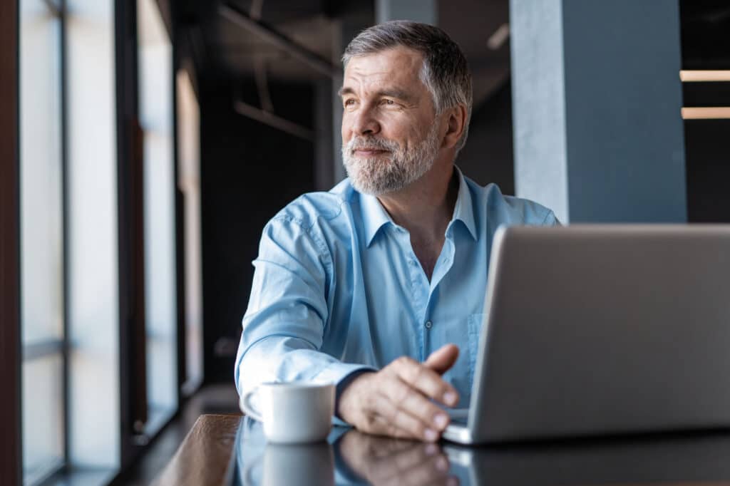 Middle-aged man with gray beard sitting at a table with laptop and coffee cup, looking out window.