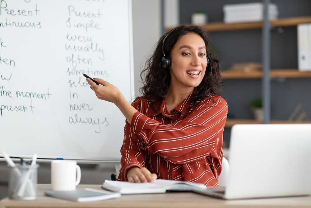 Woman with headset teaching online in front of whiteboard with English grammar notes.