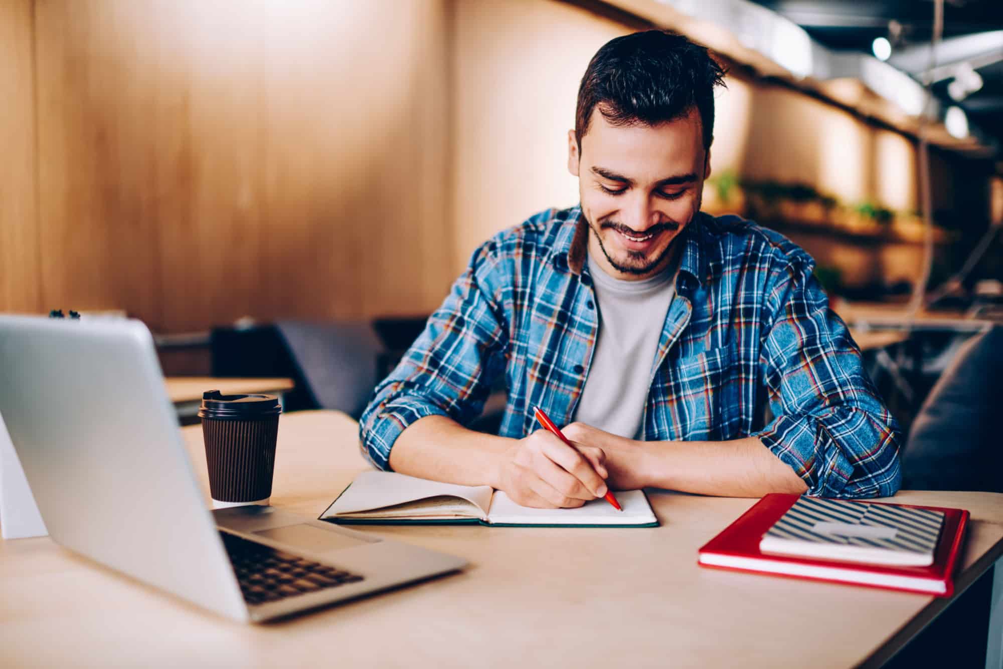 Smiling man in plaid shirt writing in notebook at table with laptop and coffee cup nearby.