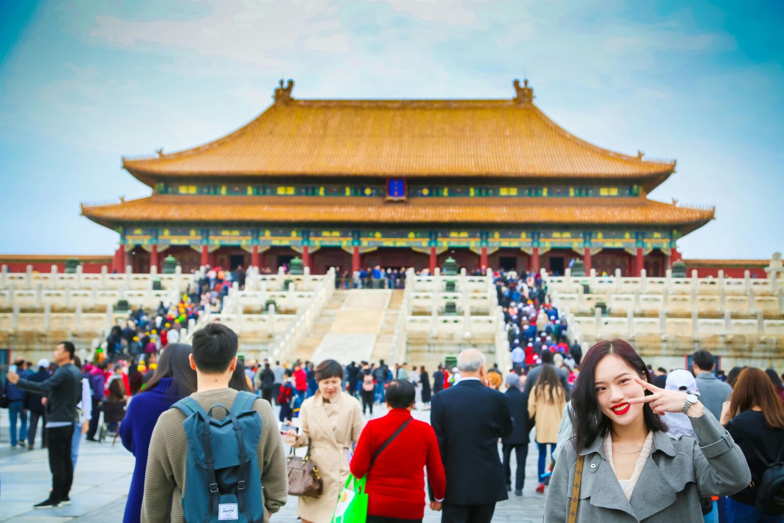 Woman in gray coat smiling and posing with a peace sign near a crowded traditional Chinese palace.
