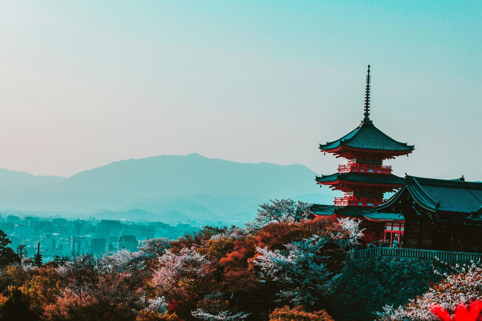 Traditional Japanese pagoda above colorful trees with cityscape and mountains in the background.