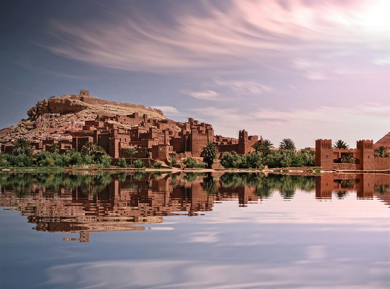 Ancient desert fortress village with palm trees reflected in calm water under a pinkish sky.