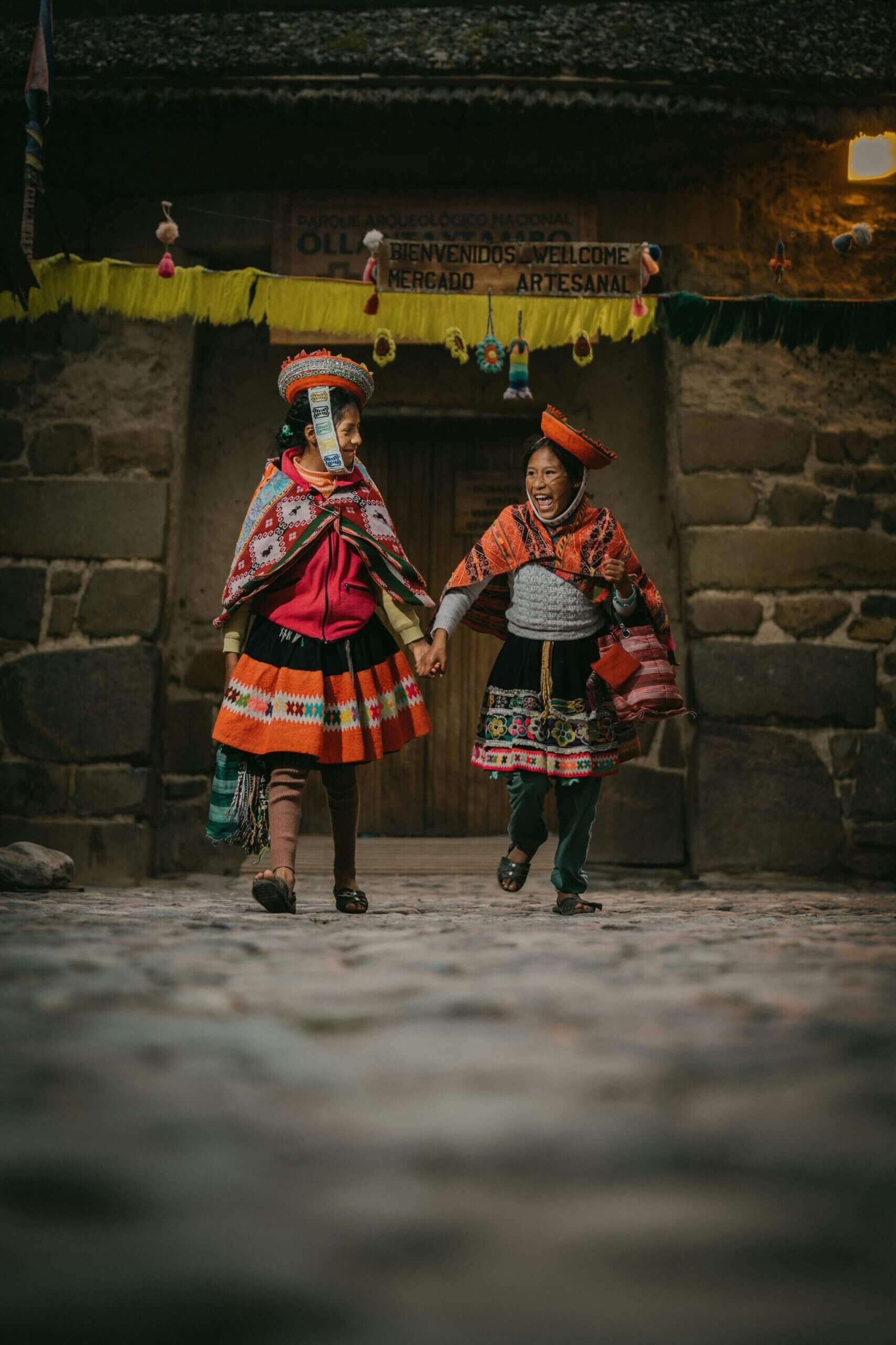 Two women in colorful traditional clothing holding hands and walking on a cobblestone street.