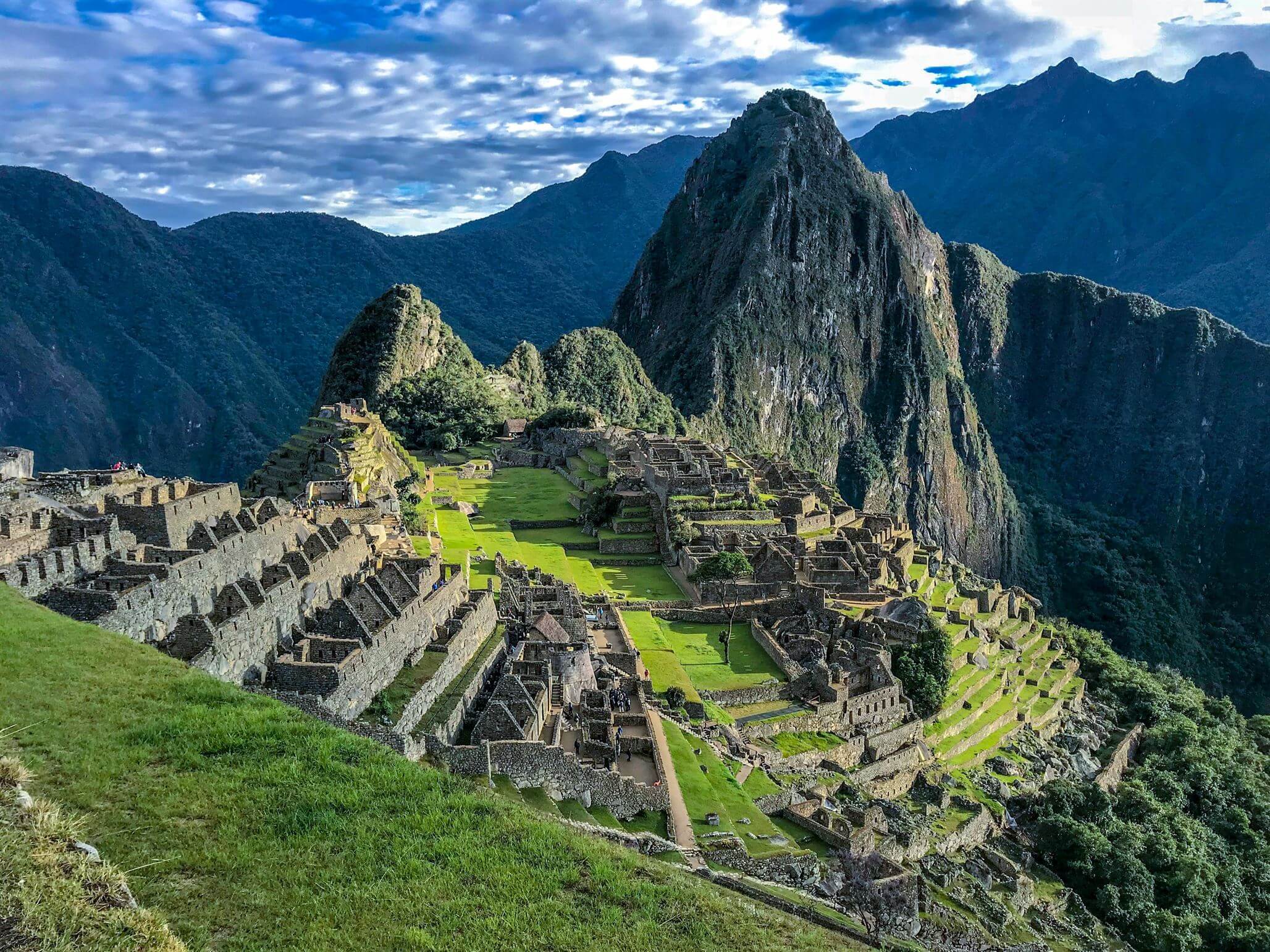 Ancient stone ruins of Machu Picchu with green terraces and surrounding mountains under a cloudy sky.