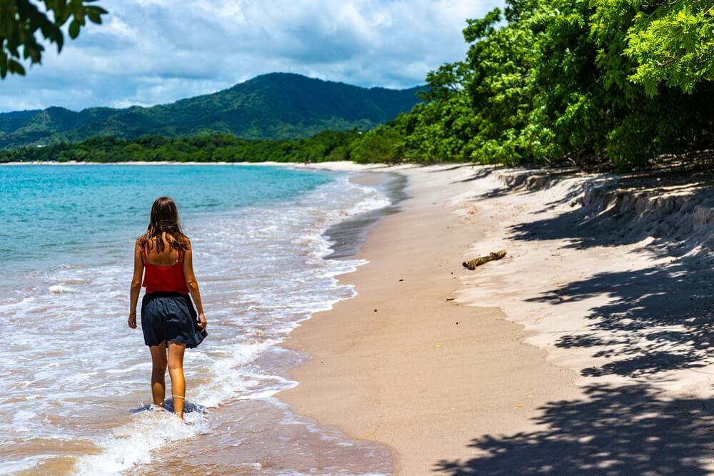 Woman walking barefoot along a sandy beach with turquoise water and forested mountains in the background.