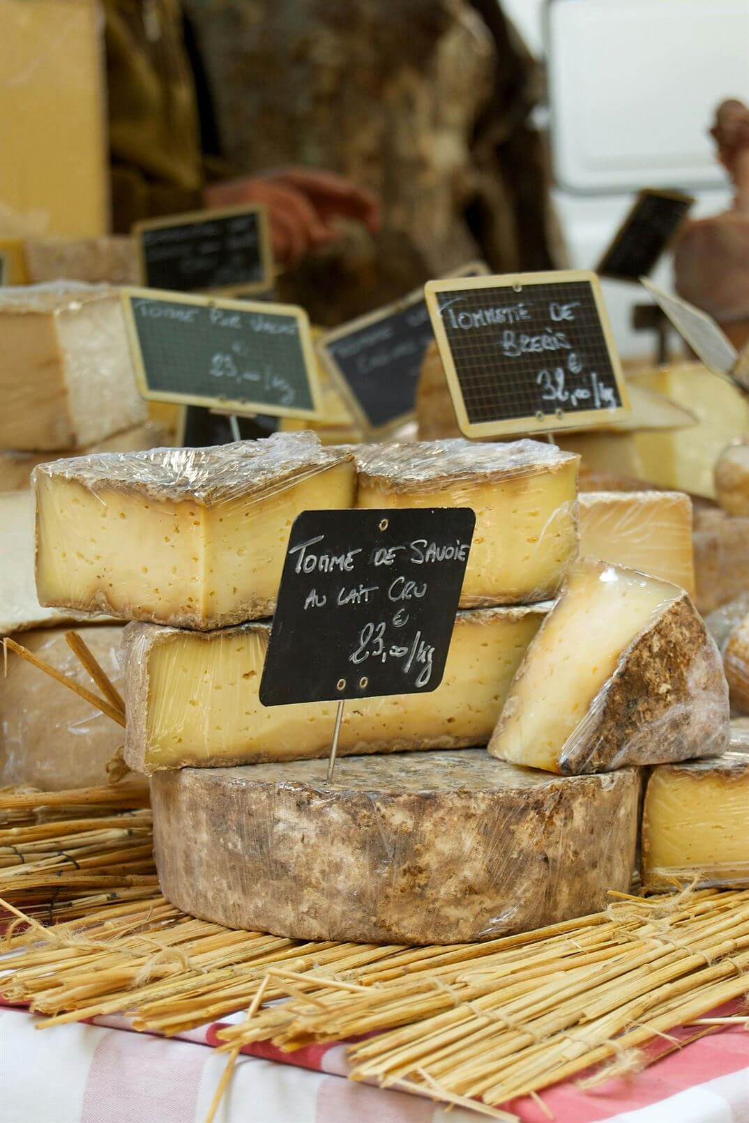 Various cheeses on display with labels showing types and prices in a rustic market setting.