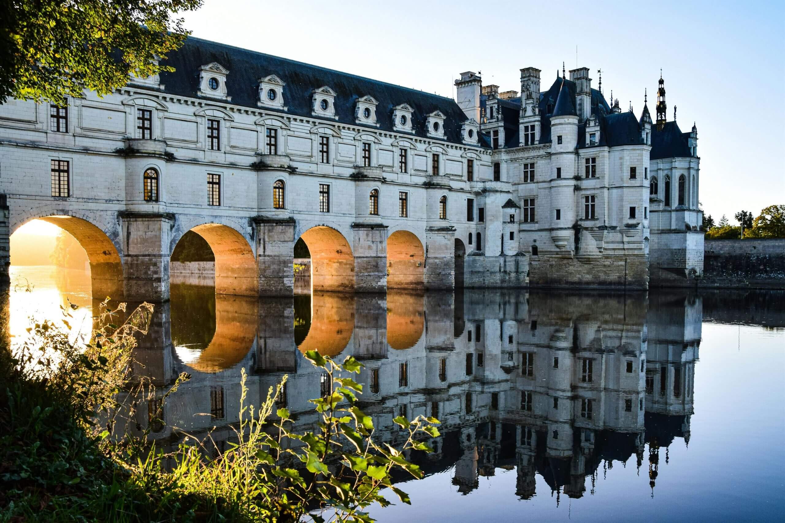 Historic stone castle with arched bridge over calm river reflecting the building and arched windows.