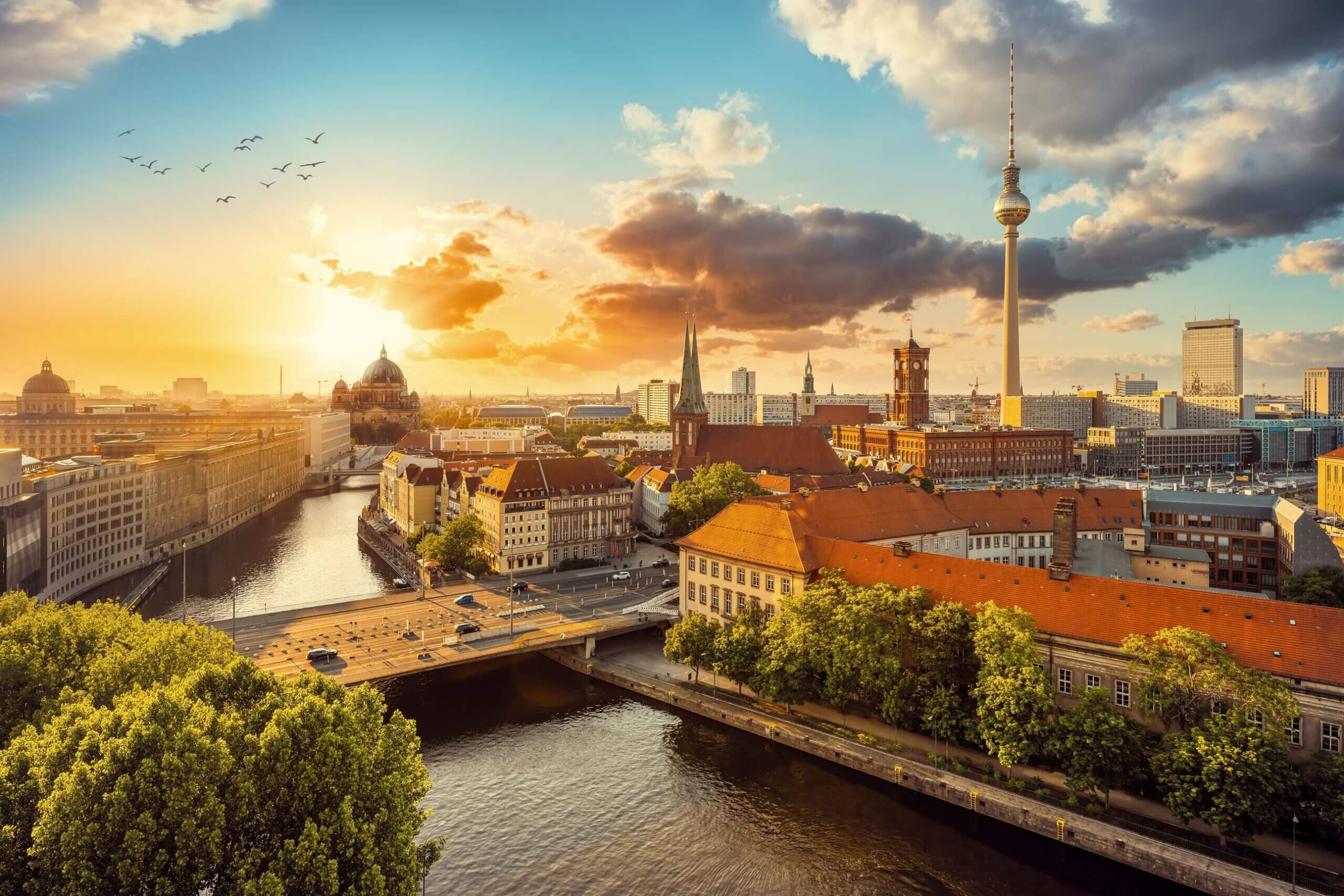 Sunset over Berlin with river, bridges, TV Tower, historic buildings, and birds flying in sky.